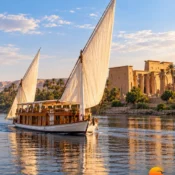 Traditional two-masted dahabiya with white sails and a shaded wooden upper deck sailing on the Nile beside an ancient temple
